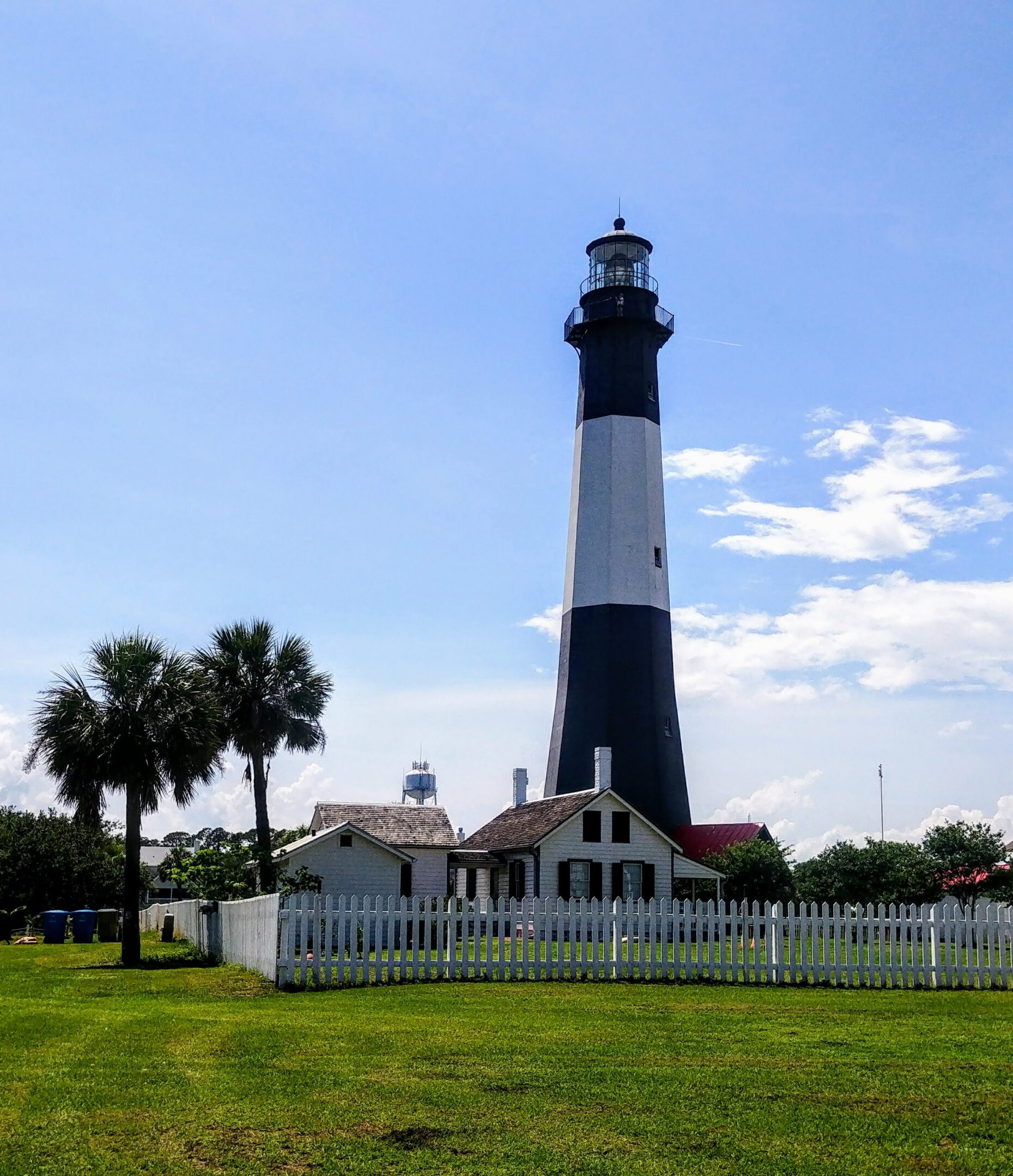 Striped lighthouse with surrounding fence and trees.